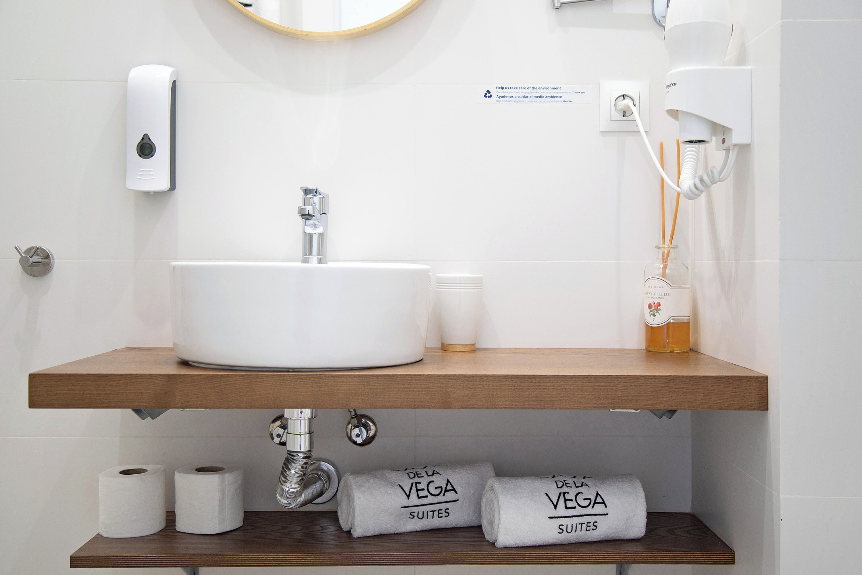 A bathroom with a white sink, a mirror, a hand soap dispenser, a hairdryer, and two towels labeled "DE LA VEGA SUITES".
