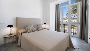 Une chambre dotée d’un grand lit, de deux tables de chevet, d’une commode et offrant une vue sur les palmiers et les immeubles grâce à de grandes fenêtres.