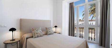 A bedroom with a large bed, two bedside tables, a nightstand, and a view of palm trees and buildings through large windows.