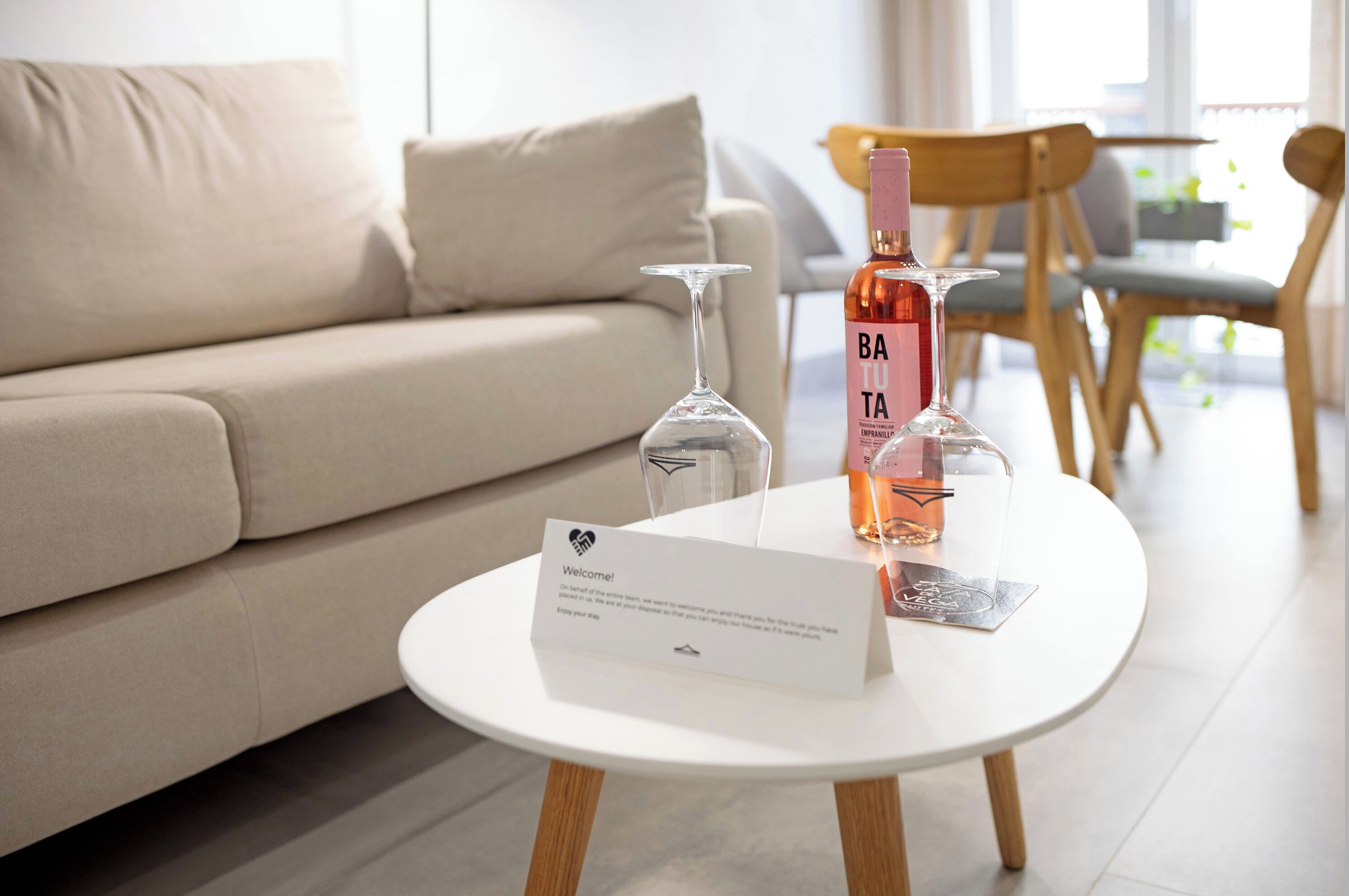 Una sala de estar moderna con un sofá color beige, una mesa redonda blanca con un letrero de bienvenida y una botella de vino.