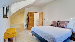 Une chambre à coucher comprenant un lit, un banc, une armoire en bois et une porte donnant sur une autre pièce.