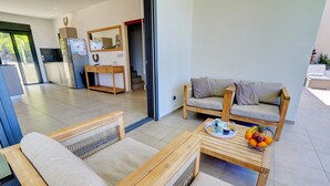 Un salon moderne avec un ensemble de canapés, une table basse sur laquelle on peut voir des fruits, et un coin cuisine visible à travers une porte ouverte.