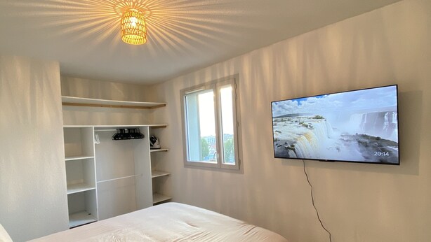 A bedroom with a bed, a TV mounted on the wall, a ceiling light, and a window. - Appartement + Parking Privé Gratuit (Marseille)