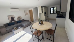 Ein modernes Wohnzimmer mit einem grauen Ecksofa, einem runden Holzesstisch mit vier Stühlen und einer Küchenzeile inklusive Waschmaschine und Fenster.