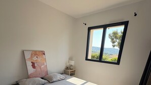 Ein Schlafzimmer mit Bett, Kissen, Nachttisch, Lampe, Fenster mit Aussicht und einem Wandbild.