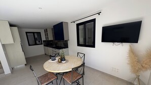 Eine moderne Küche mit dunklen Arbeitsplatten, weißen Schränken und einer blauen Spritzschutzwand. Ein Essbereich mit einem runden Tisch und zwei Stühlen, ein an der Wand befestigtes Fernsehgerät und eine Vase mit Trockenpflanzen auf dem Boden.