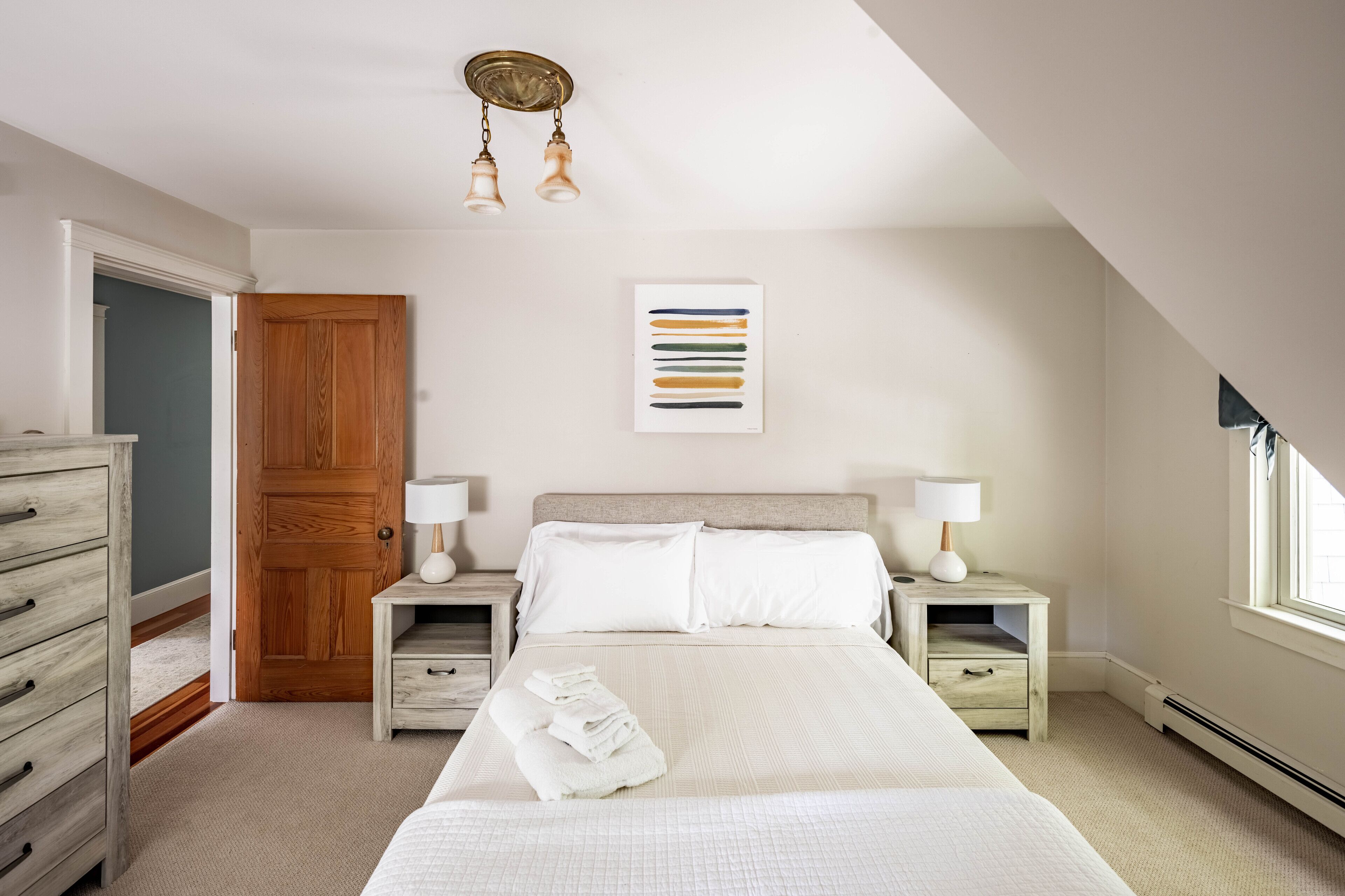 A bedroom with a large bed, two bedside tables with lamps, a wooden door, and a framed artwork on the wall.
