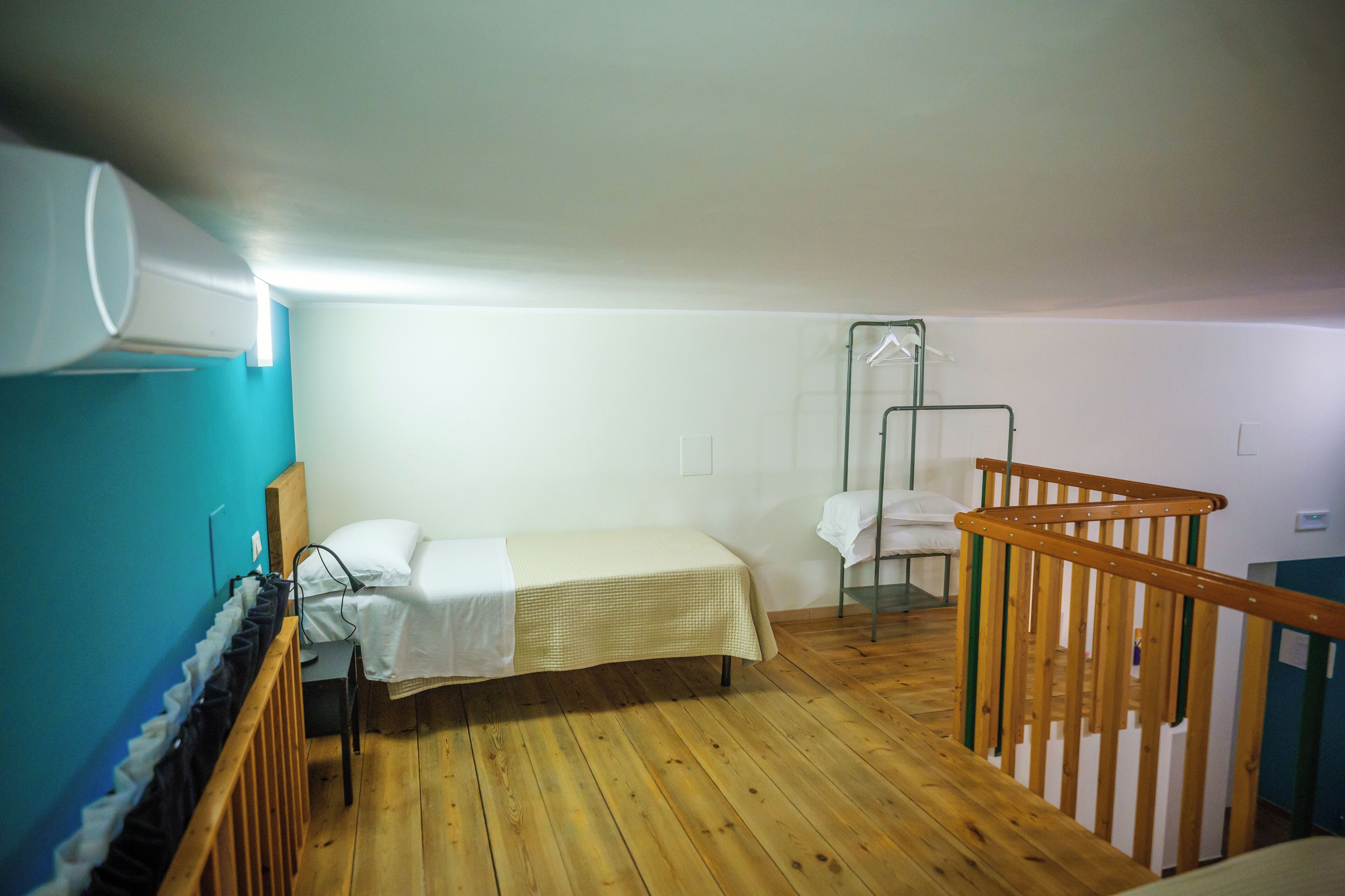 Een kleine eenpersoonskamer met een houten vloer, een wit bed, een houten leuning en een kledingrek.