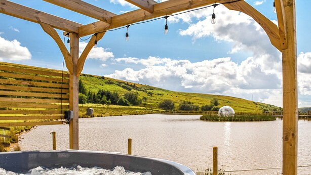 Ein Whirlpool unter einer Holzpergola mit Blick auf einen See und Hügel.