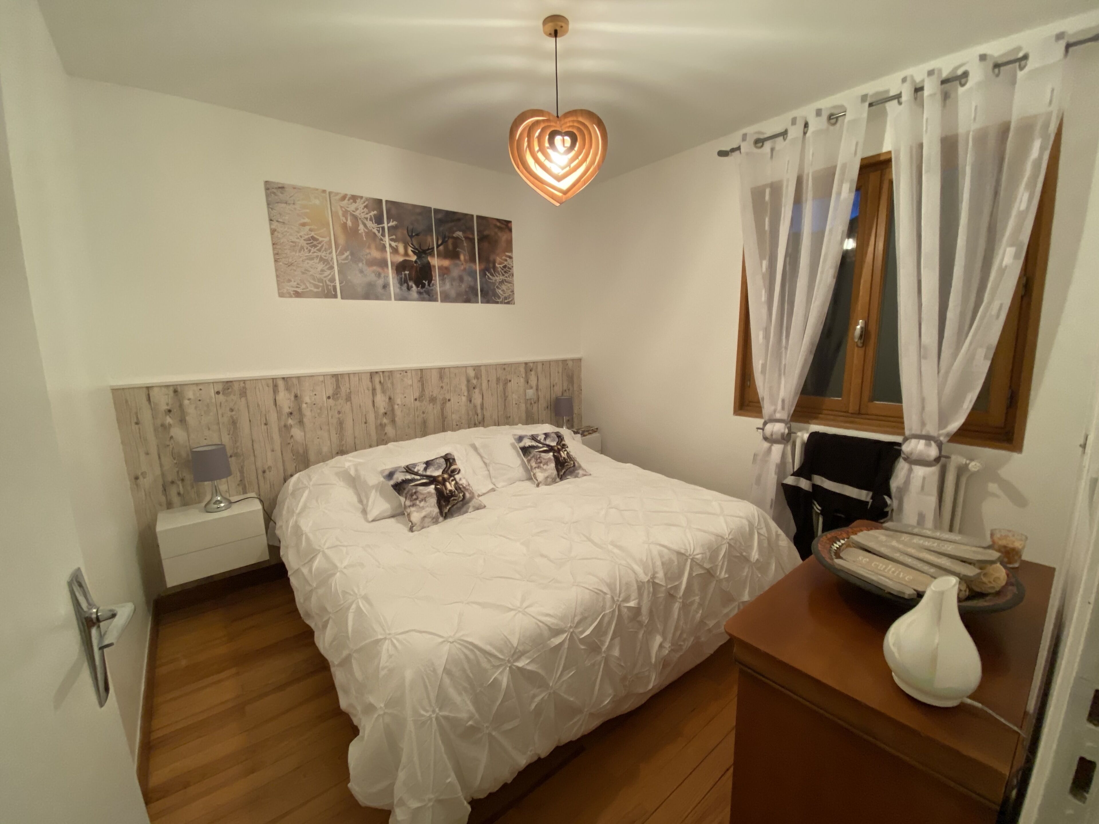 A bedroom with a wooden headboard, a bed with white bedding, a nightstand with a lamp, a wall-mounted artwork, and a bedside table with a vase and books.
