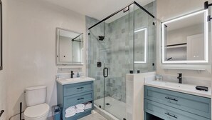 A modern bathroom with a glass-enclosed shower, a white toilet, and two vanity areas with blue cabinets. - Coastal studio w/ beach access & free parking (San Clemente)