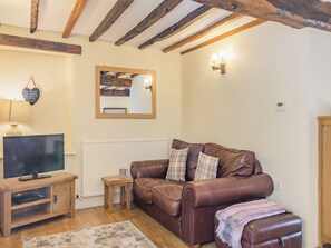 A cozy living room with a brown leather sofa, a wooden TV stand, a small side table, a mirror, and a wall-mounted light fixture. - Bluebell Cottage - UKC6676 (Betws Yn Rhos)