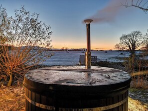 A large, cylindrical outdoor heater with a chimney emitting smoke, surrounded by a snowy landscape and trees.