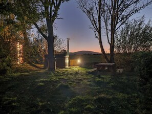 A rustic outdoor setting with a wooden bench, a metal structure, and a pathway leading to a building with a lit window. - Bluebell Cottage - UKC6676 (Betws Yn Rhos)