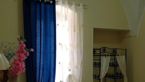 A room with a tiled floor, a wooden side table with a vase of pink flowers, a window with blue and white curtains, and a metal rack with white curtains.