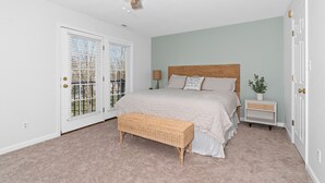 A bedroom with a large bed, a ceiling fan, a bench, a nightstand, and a window with blinds.
