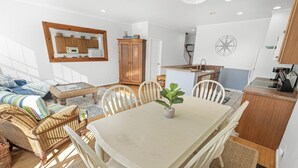 A spacious living area with a dining table, white chairs, wicker furniture, and a kitchen area with wooden cabinets and a white countertop.