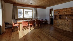 Tilava huone, jossa on puulattia, ruokailutila pöytineen ja tuoleineen, ikkuna verhoilla sekä puinen kaappi laatikoineen.
