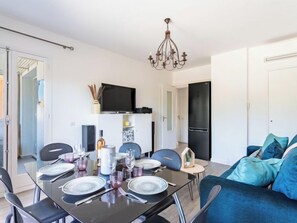 A modern living room with a dining area, a black sofa, teal cushions, a chandelier, and a flat-screen TV mounted on the wall. - Mer et Soleil I by Interhome (Bormes-les-Mimosas)