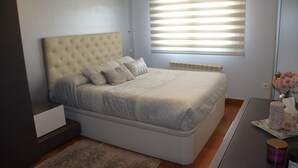 Une chambre avec un grand lit, une table de chevet, une commode, une fenêtre avec des stores et une petite table sur laquelle se trouvent un vase et des bougies.