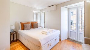 Un dormitorio con una cama grande, una mesita de noche, una lámpara y una ventana con vistas a edificios.