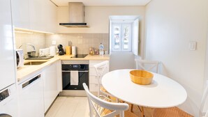 Cocina moderna con armarios blancos, horno empotrado, microondas y cafetera. Hay una mesa redonda con un cuenco de madera y sillas blancas.