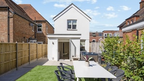 Ein modernes, zweistöckiges Haus mit weißer Fassade, einer Terrasse mit Tisch und Stühlen sowie einem kleinen Garten.