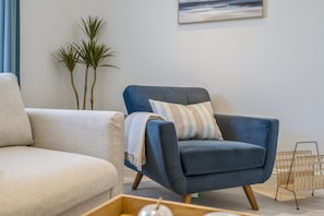 Een moderne woonkamer met een blauwe fauteuil, een beige bank en een kleine houten tafel met decoratieve objecten.