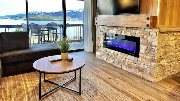 A modern living room with a stone fireplace, a flat-screen TV mounted above it, a comfortable sofa, and a view of a lake and mountains through large sliding doors. - Cozy, Modern Lakeside Condo (Dillon)