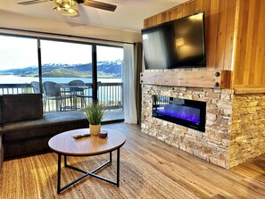 A modern living room with a stone fireplace, a flat-screen TV mounted above it, a comfortable sofa, and a view of a lake and mountains through large sliding doors. - Cozy, Modern Lakeside Condo (Dillon)