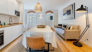A modern kitchen with white cabinets, a dining table with a patterned tablecloth, and a brown chair. There is a sofa and a floor lamp in the living area. - Portuguese Designed Apartment with Terase (Lisboa)