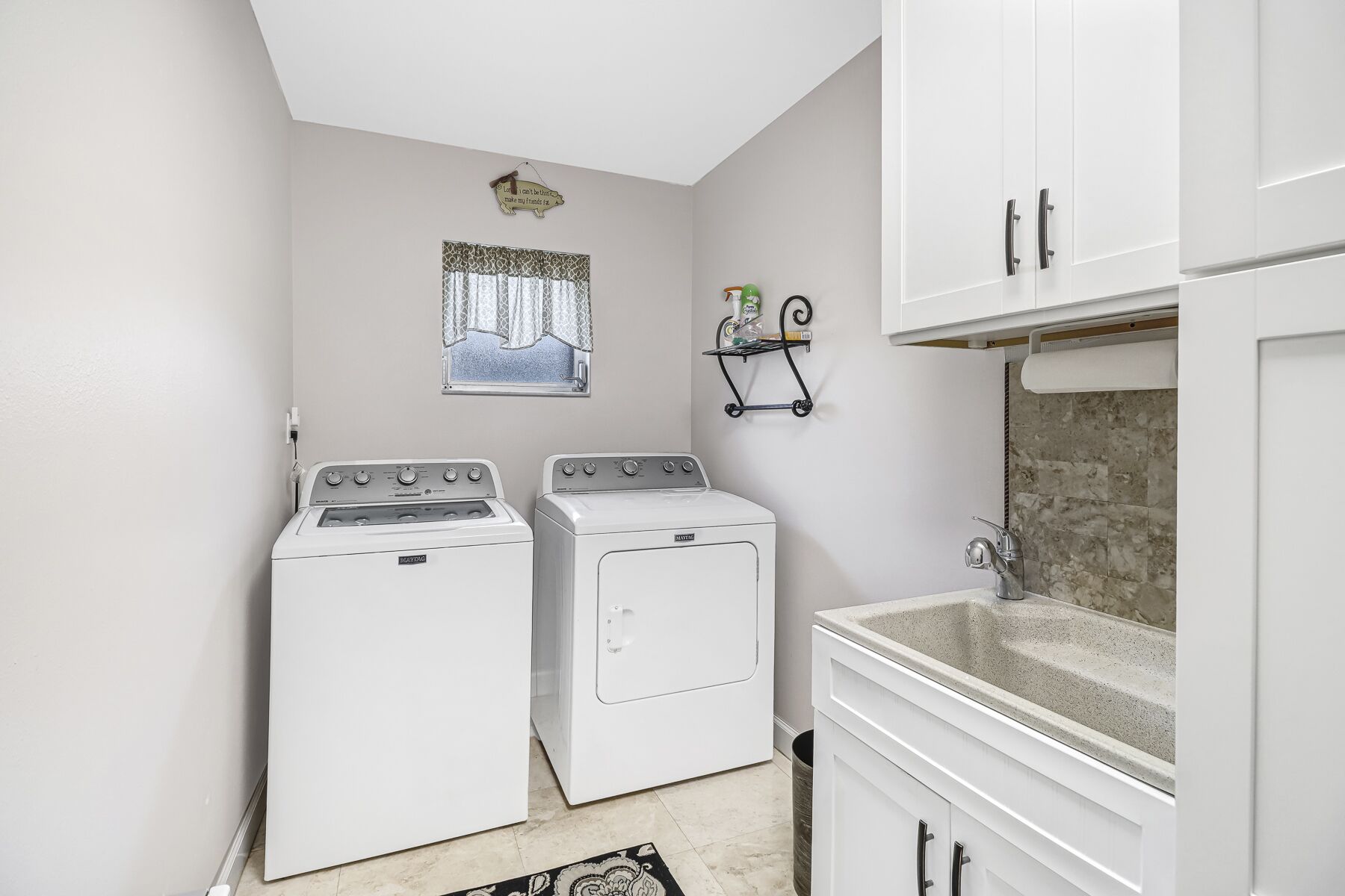 A laundry room with two front-loading washers and dryers, a small window with curtains, a wall-mounted shelf with toiletries, and a sink with a marble countertop.