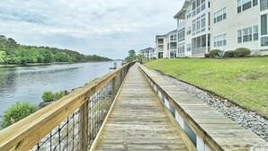 A wooden boardwalk leading to a white building with multiple windows and balconies, overlooking a river.