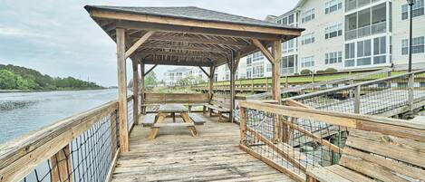 A wooden pier with a covered seating area and picnic tables, overlooking a body of water.