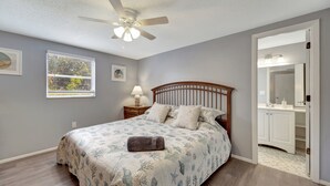 A bedroom with a bed, a ceiling fan, a window, a nightstand, and a bathroom with a mirror and cabinets.