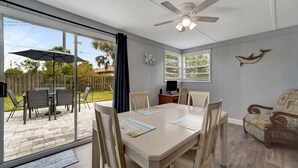 A room with a large sliding glass door leading to an outdoor patio, a ceiling fan, a dining table with chairs, and a television on a stand.