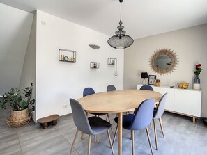 Uma sala de jantar com uma mesa de madeira redonda, seis cadeiras azuis, um lustre preto pendurado, um aparador branco com um espelho e um vaso de planta.