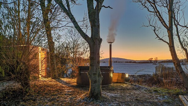 A rustic outdoor setting with a wooden structure, a chimney emitting smoke, and a frozen lake in the background. - Primrose Cottage - UKC6679 (Betws Yn Rhos)