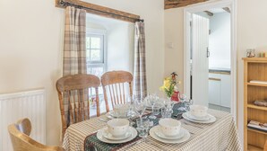 A dining area with a table set for four, wooden chairs, a window with curtains, and a view into another room. - Primrose Cottage - UKC6679 (Betws Yn Rhos)