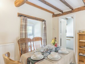 A dining area with a table set for four, wooden chairs, a window with curtains, and a view into another room. - Primrose Cottage - UKC6679 (Betws Yn Rhos)