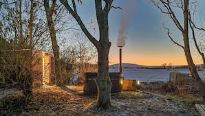 A rustic outdoor setting with a wooden structure, a chimney emitting smoke, and a frozen lake in the background. - Lavender Cottage - UKC6677 (Betws Yn Rhos)
