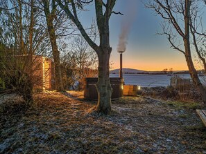 A rustic outdoor setting with a wooden structure, a chimney emitting smoke, and a frozen lake in the background. - Lavender Cottage - UKC6677 (Betws Yn Rhos)