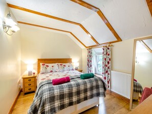 A bedroom with a wooden ceiling, a bed with floral bedding, a nightstand with a lamp, and a window with curtains.