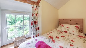 A bedroom with a wooden headboard, a large window with curtains, a wicker basket, and a floral bedspread. - Lavender Cottage - UKC6677 (Betws Yn Rhos)