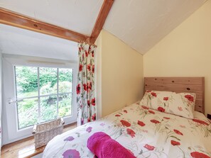 A bedroom with a wooden headboard, a large window with curtains, a wicker basket, and a floral bedspread. - Lavender Cottage - UKC6677 (Betws Yn Rhos)