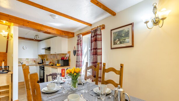 A dining area with a table set for four, including plates, glasses, and a silver pitcher. A wooden chair is positioned at the table, and a kitchen is visible in the background. - Lavender Cottage - UKC6677 (Betws Yn Rhos)