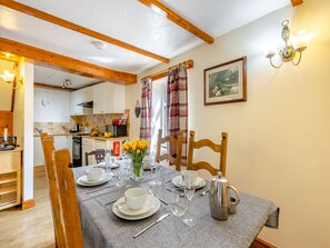 A dining area with a table set for four, including plates, glasses, and a silver pitcher. A wooden chair is positioned at the table, and a kitchen is visible in the background. - Lavender Cottage - UKC6677 (Betws Yn Rhos)