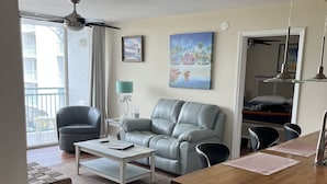 Een zithoek met een grijze leren bank, een witte salontafel, en een eethoek met een houten tafel en stoelen. Aan de muur hangt een schilderij en erboven de bank hangt een plafondventilator.