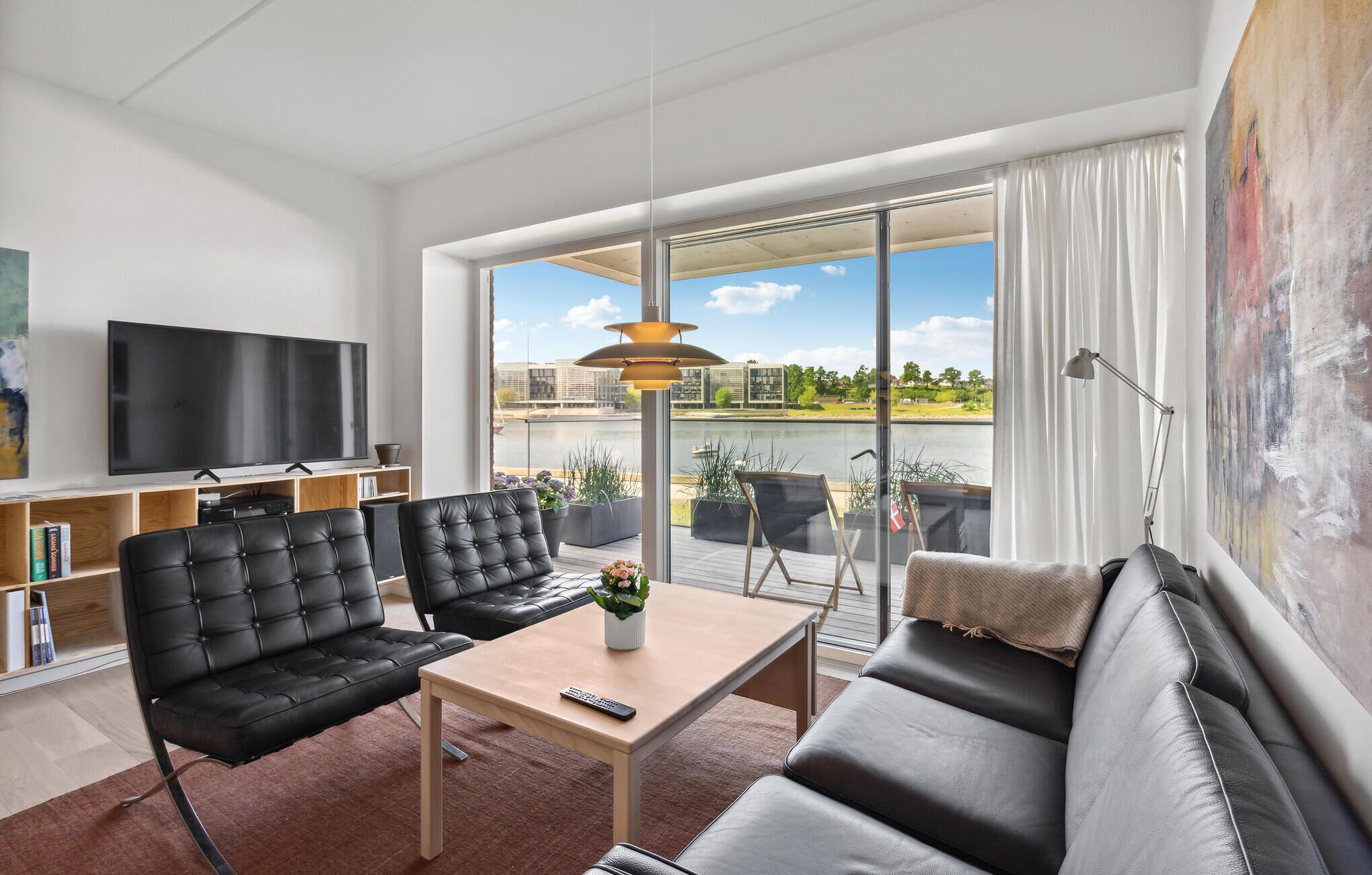 Un salon moderne doté d’un grand téléviseur, de sièges confortables et offrant une vue sur une rivière et des bâtiments.