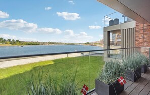 Un balcon moderne avec une balustrade en verre donnant sur une rivière, avec une pelouse et des plantes en pot au premier plan.